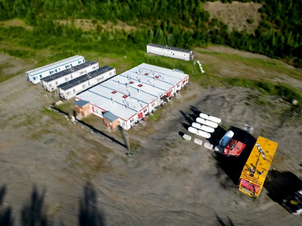 Aerial view of a remote site with 1998 / Relocatable Industrial Accommodation—rectangular buildings, trailers, propane tanks, and trucks on a gravel lot—surrounded by dense green forest.