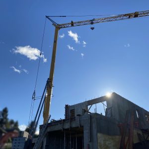 The 2024 XJCM Self Erecting Crane stands tall over a partially built concrete structure under a clear blue sky, sunlight shining behind. Various construction materials and equipment are visible on the site.