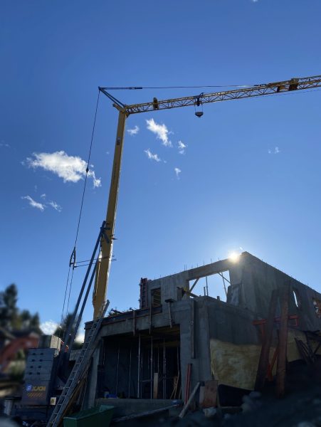 The 2024 XJCM Self Erecting Crane stands tall over a partially built concrete structure under a clear blue sky, sunlight shining behind. Various construction materials and equipment are visible on the site.