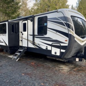 A modern travel trailer with black, white, and gray stripes is parked on a gravel area beside trees. The trailer has several windows, an extended slide-out section, and entrance steps deployed.