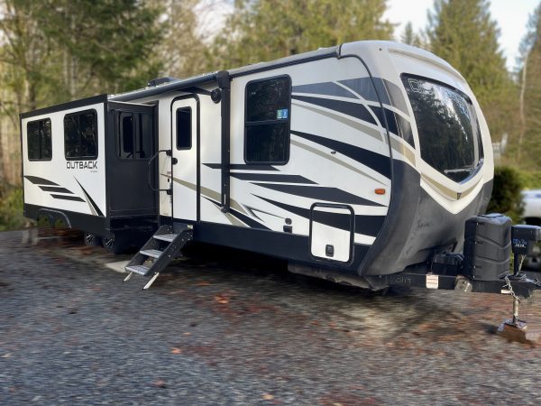 A modern travel trailer with black, white, and gray stripes is parked on a gravel area beside trees. The trailer has several windows, an extended slide-out section, and entrance steps deployed.