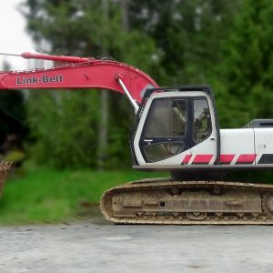 A red and white 2006 Link-Belt 200SH Excavator sits on concrete with its arm extended, surrounded by greenery and trees in the background.