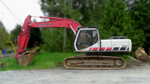 A red and white 2006 Link-Belt 200SH Excavator sits on concrete with its arm extended, surrounded by greenery and trees in the background.