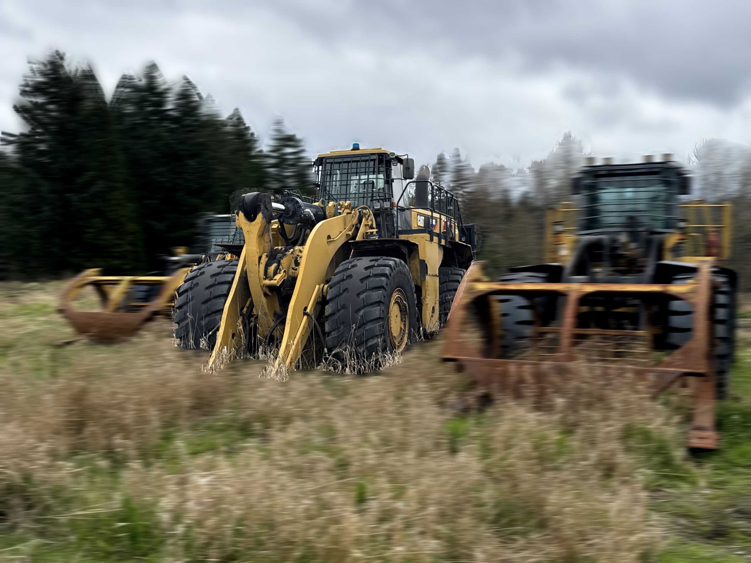 A 2017 / 988K CAT Front End Loader with large tires is parked in a grassy field among trees and similar equipment, beneath a cloudy sky.