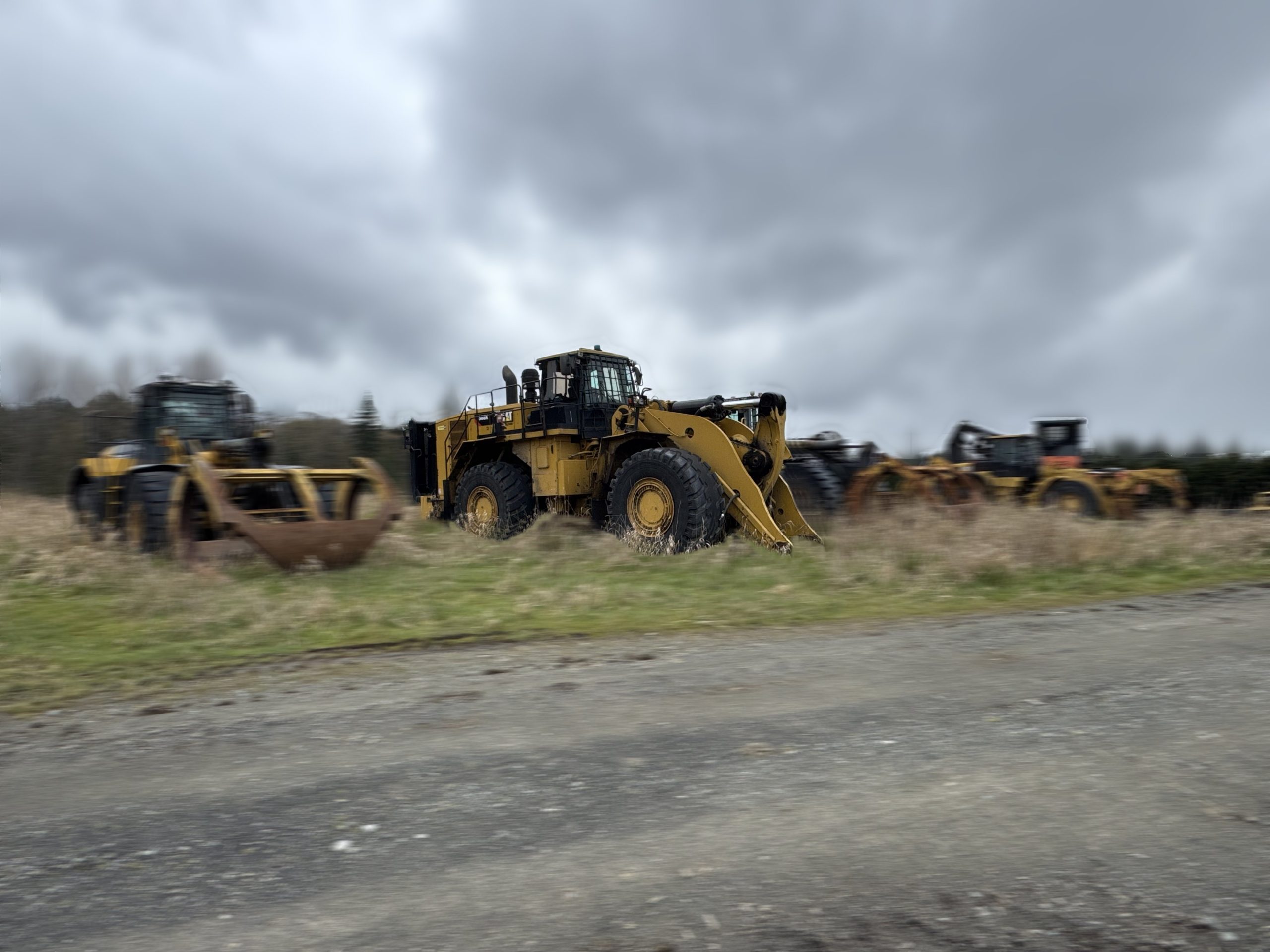 A group of large yellow construction vehicles, including a 2017 / 988K CAT Front End Loader and bulldozers, are parked on a grassy field under a cloudy sky, with a gravel road running in front of them.