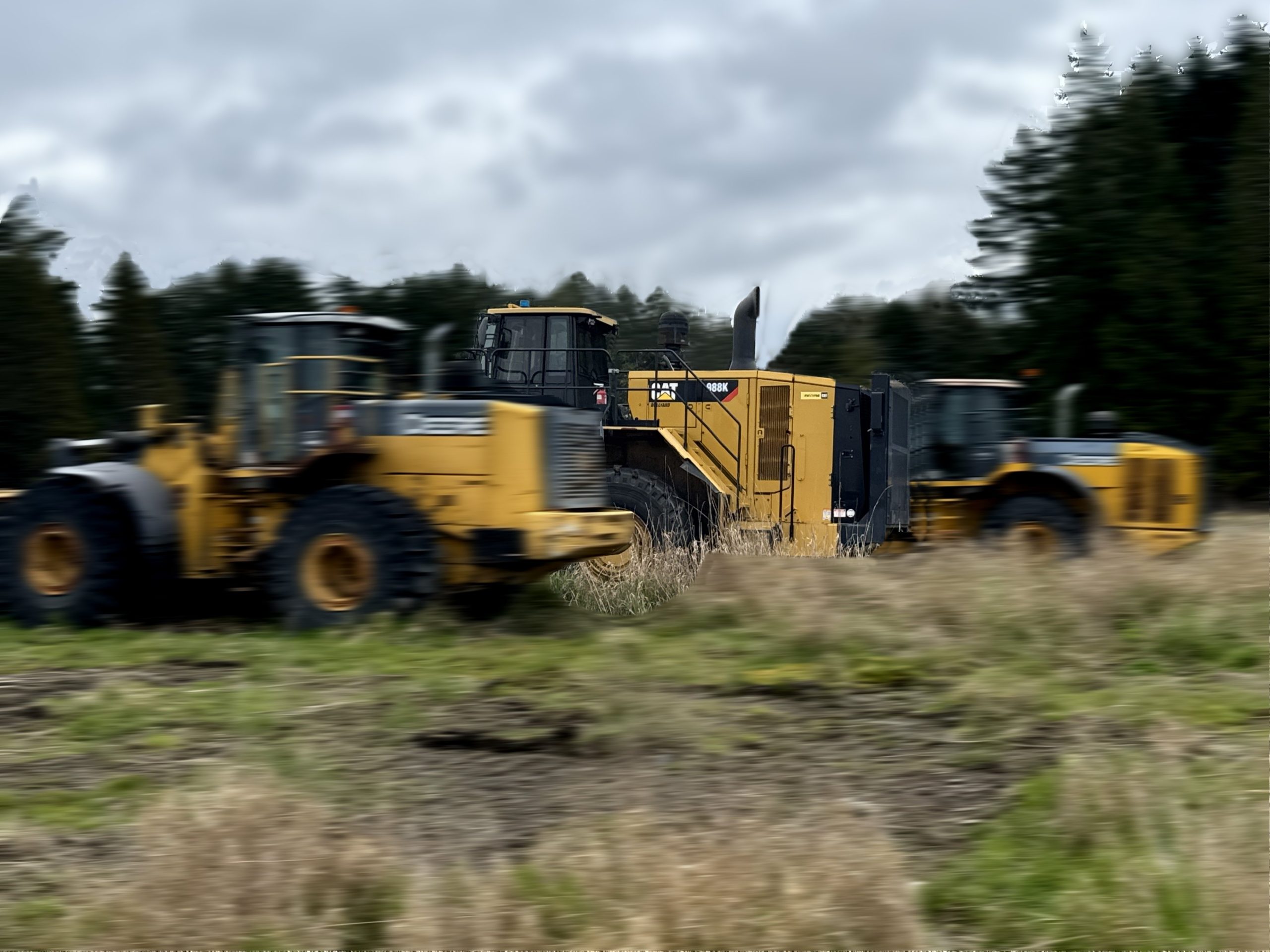 Three large yellow construction vehicles, including a 2017 / 988K CAT Front End Loader and an industrial grinder, are parked on a grassy field with trees and a cloudy sky in the background. The image is slightly blurred.
