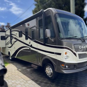 A large, modern RV with a brown and white striped design is parked on a driveway beside a lawn and trees under a partly cloudy sky.