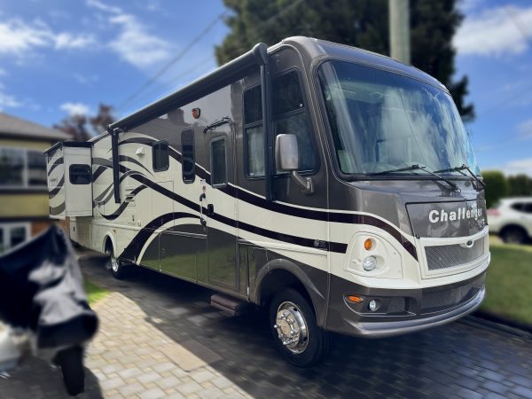 A large, modern RV with a brown and white striped design is parked on a driveway beside a lawn and trees under a partly cloudy sky.