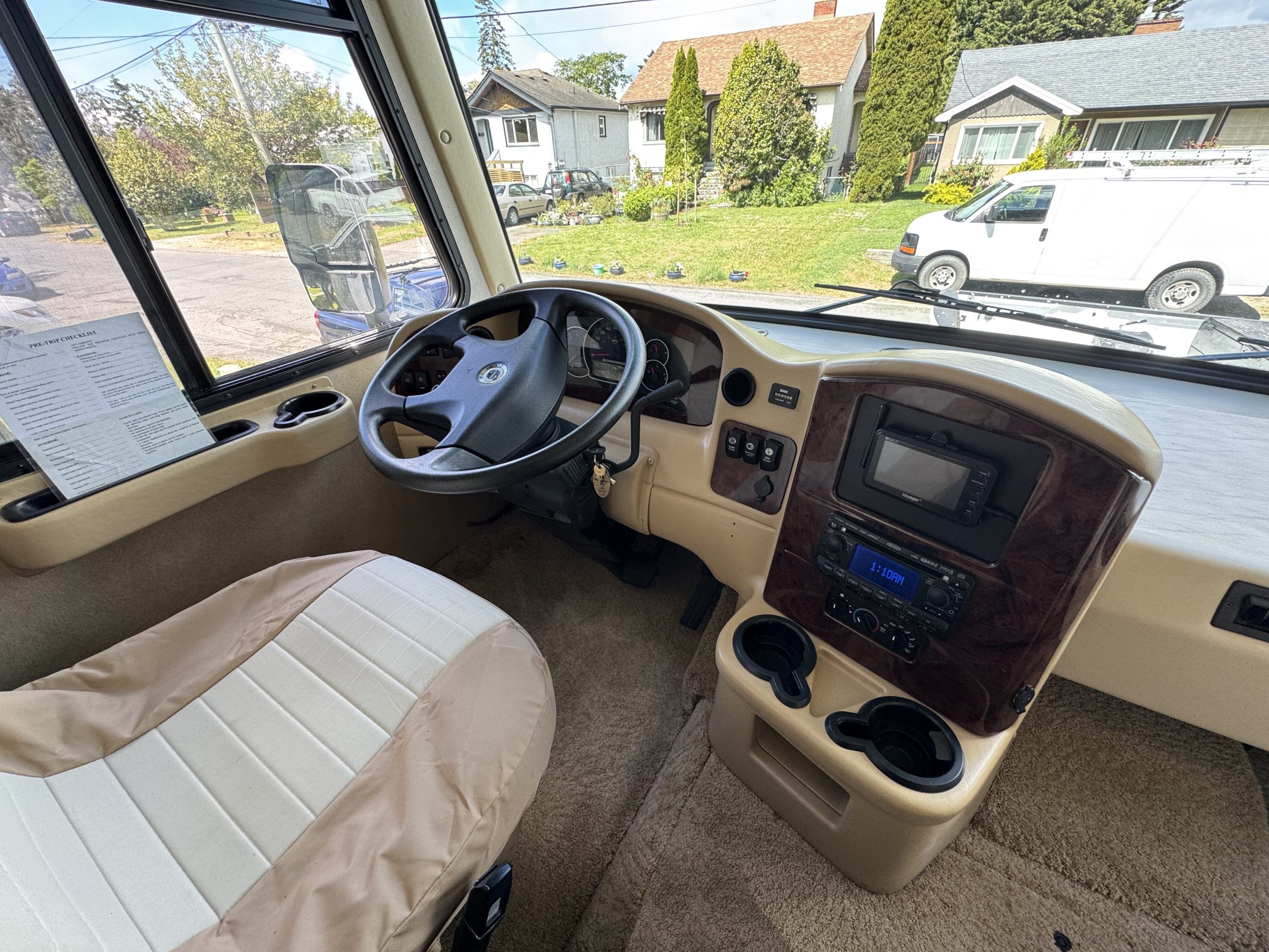 Driver’s seat and steering wheel of a 2007-10 Dumont Challenger RV with a beige interior, dashboard controls, cup holders, and display screen. Large windows reveal a suburban street with houses and parked vehicles outside.