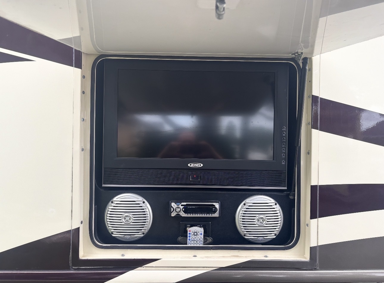 A flat-screen TV, two round speakers, a stereo system, and a remote control are mounted in an outdoor compartment of the Damon Challenger LE Model 348, a 38-foot luxury motor coach with a beige and brown exterior.