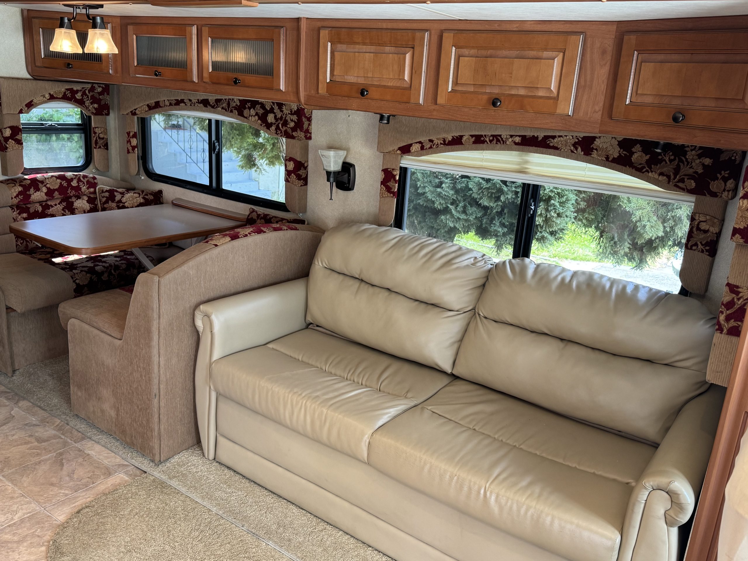 The 2007-10 Dumont Challenger features a beige leather sofa, a dining area with a wooden table and cushioned benches, overhead wooden cabinets, and windows with floral curtains that fill the interior with natural light.
