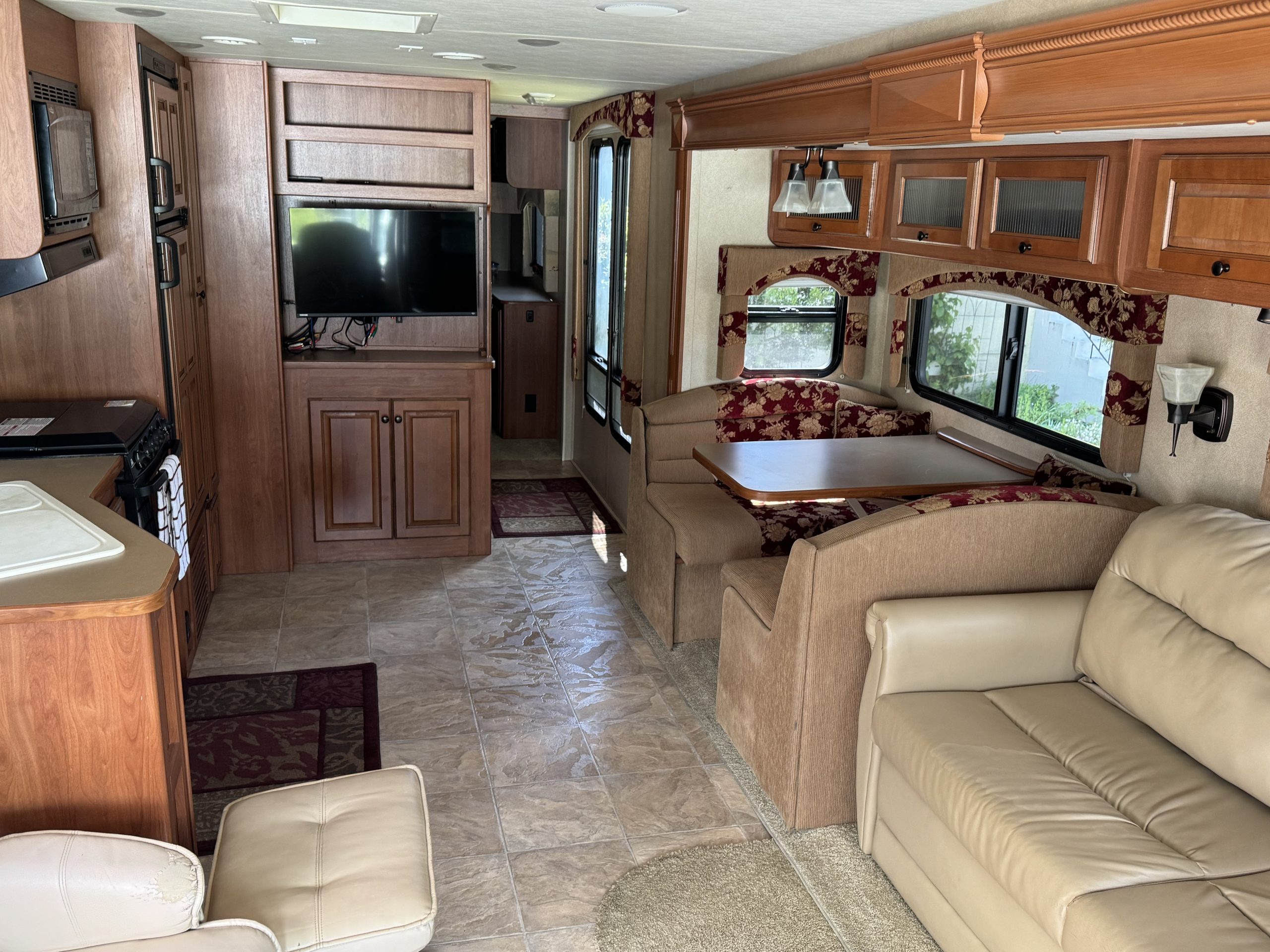 Interior of a 2007-10 Dumont Challenger features beige leather seating, cushioned dining benches, compact kitchen with wood cabinets, a mounted TV, sunlit windows, and red and beige accents for cozy decor.