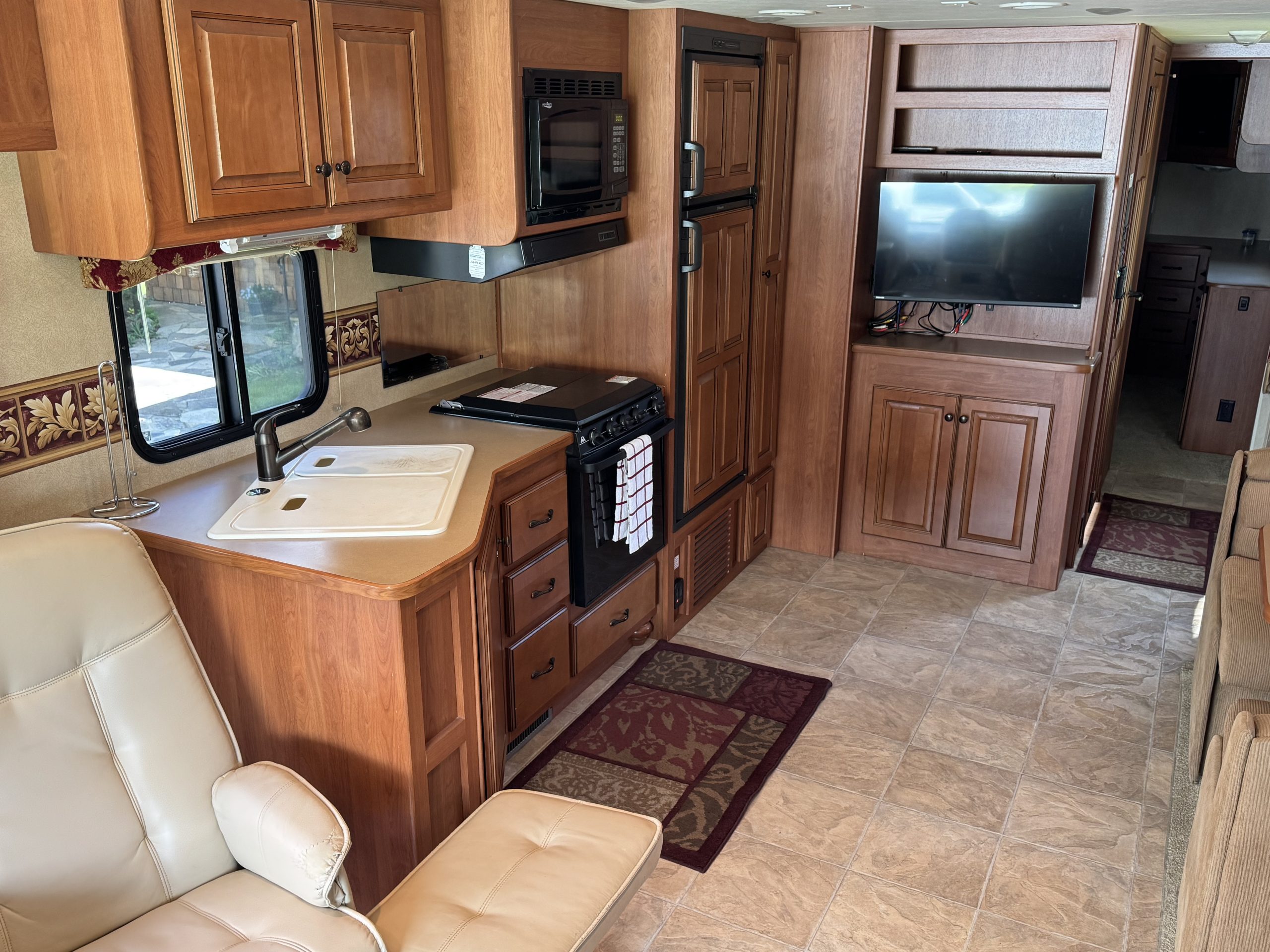 Interior of the 2007-10 Dumont Challenger RV showcasing a kitchen with sink, stove, microwave, and fridge, wooden cabinets, mounted TV, tiled floor, cream swivel chair, and patterned rugs for a cozy retreat.