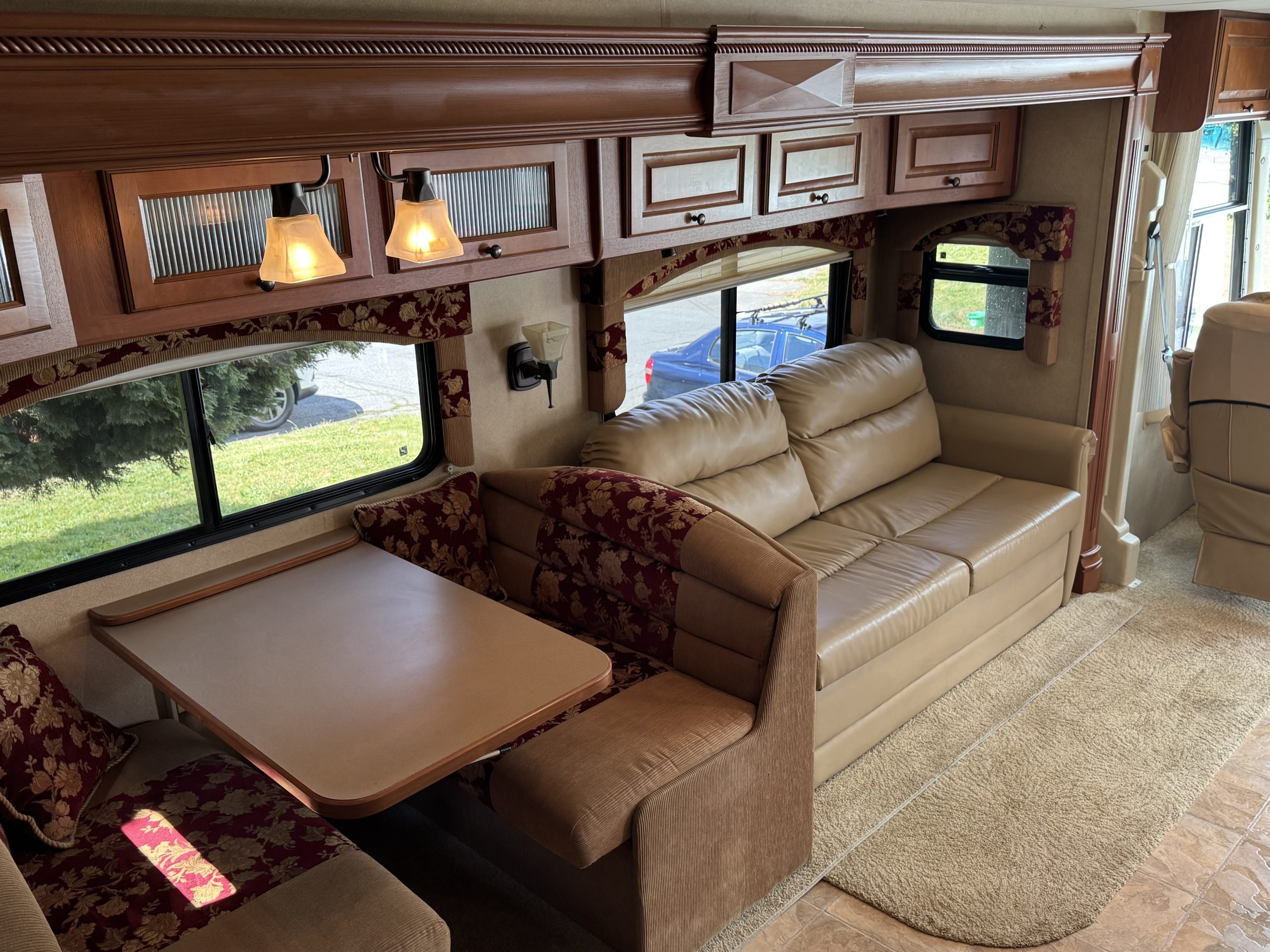 Interior of a 2007-10 Dumont Challenger featuring a dining area with table and cushioned benches, beige leather sofa, overhead wooden cabinets, windows with patterned curtains, and a carpeted floor.