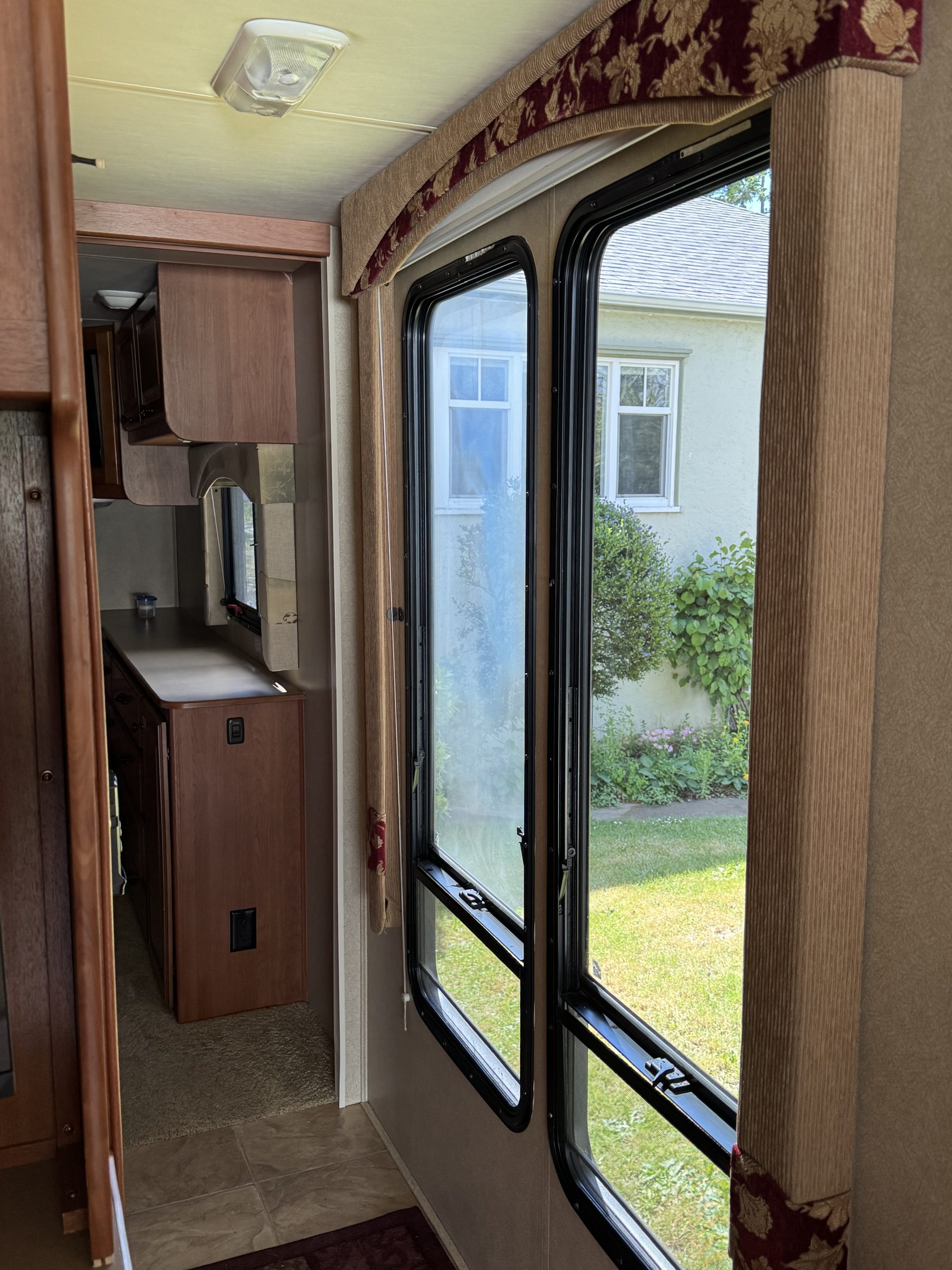 Interior view of the 2007-10 Dumont Challenger, featuring large windows with beige and maroon curtains, sunlight streaming in, a grassy outdoor view, plus wood cabinets and a compact counter for a cozy feel.