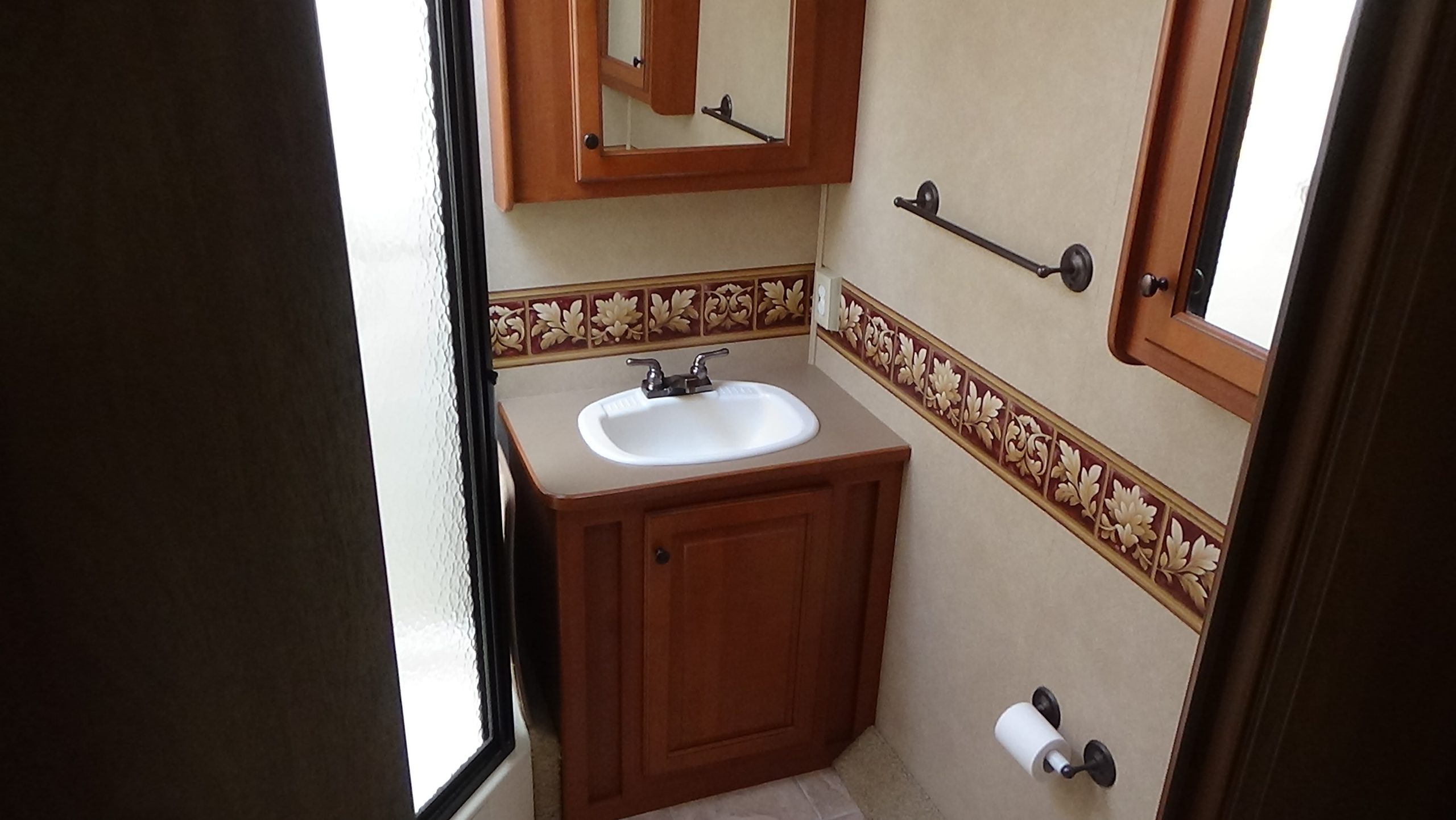Small bathroom with a single sink, wooden cabinet and mirror, floral wallpaper border, and a left glass shower door—like a serene retreat after enjoying the 2007-10 Dumont Challenger. Neutral hues on walls and floor throughout.