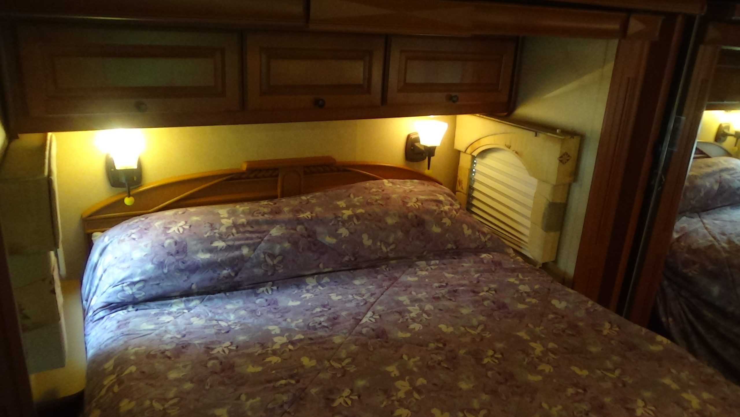 A cozy bed with a purple floral bedspread sits by a wooden headboard, built-in wall lamps, and overhead cabinets—perfect for relaxing after watching the 2007-10 Dumont Challenger—with a pleated shade covering the nearby window.