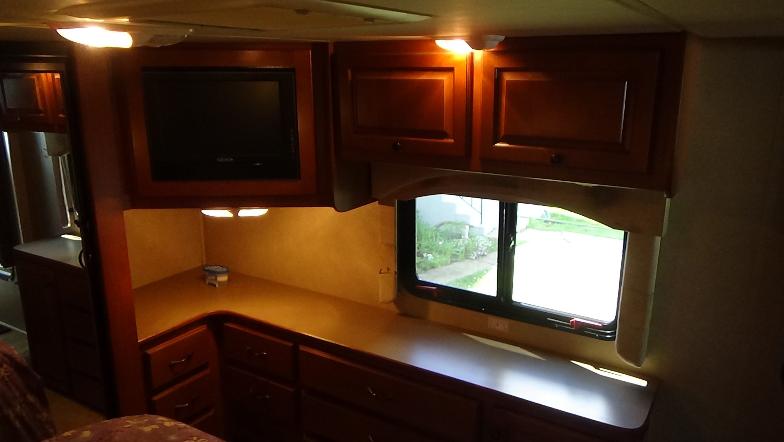 The interior of the 2007-10 Dumont Challenger RV features built-in wooden cabinets, drawers, a TV, and counter space below a sunlit window. A small round container rests on the counter.