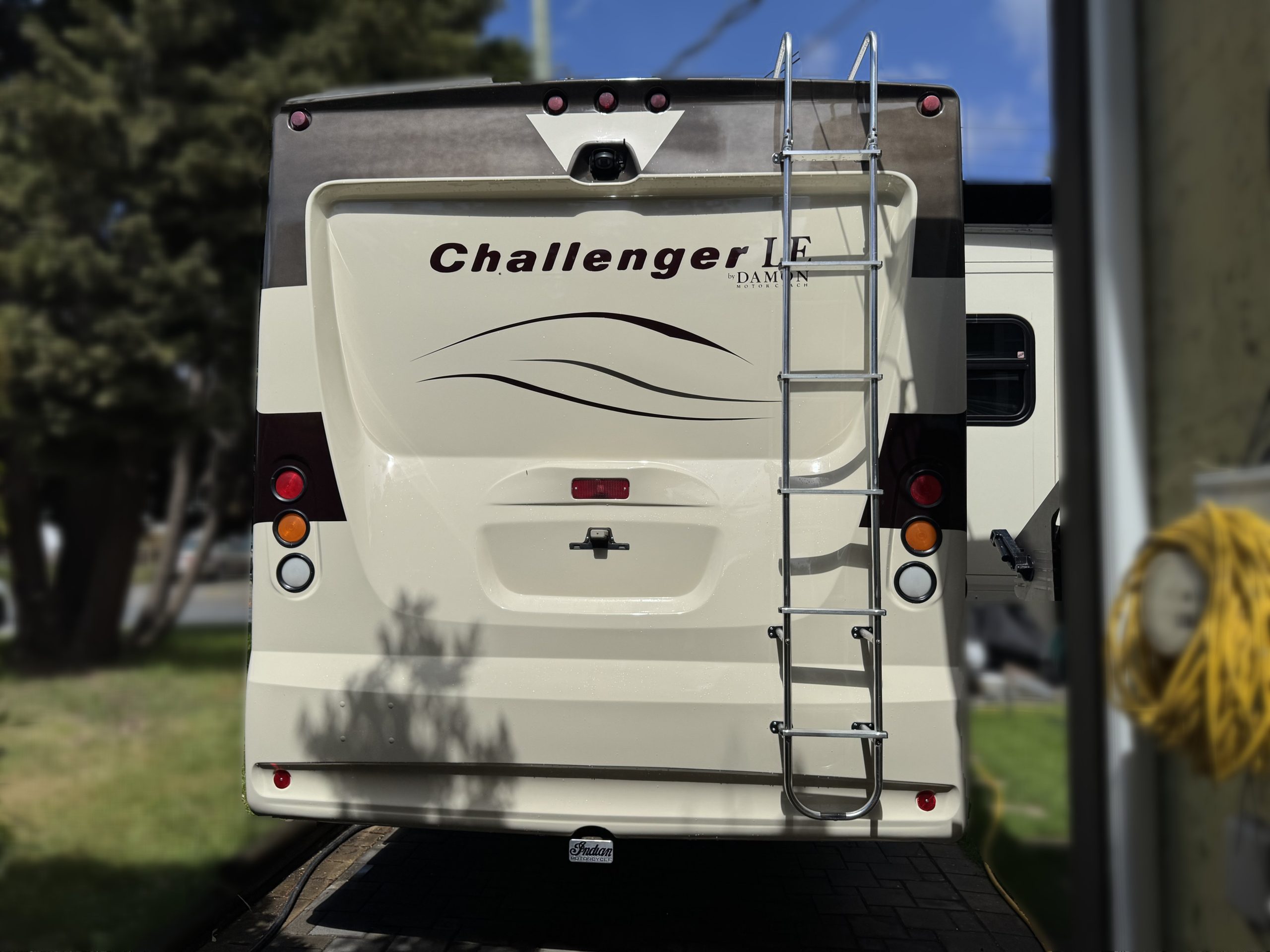 The back of a beige RV with "Challenger" written on it, featuring a rear ladder, tail lights, and subtle wave designs. Trees and a yellow coiled hose are visible in the background.