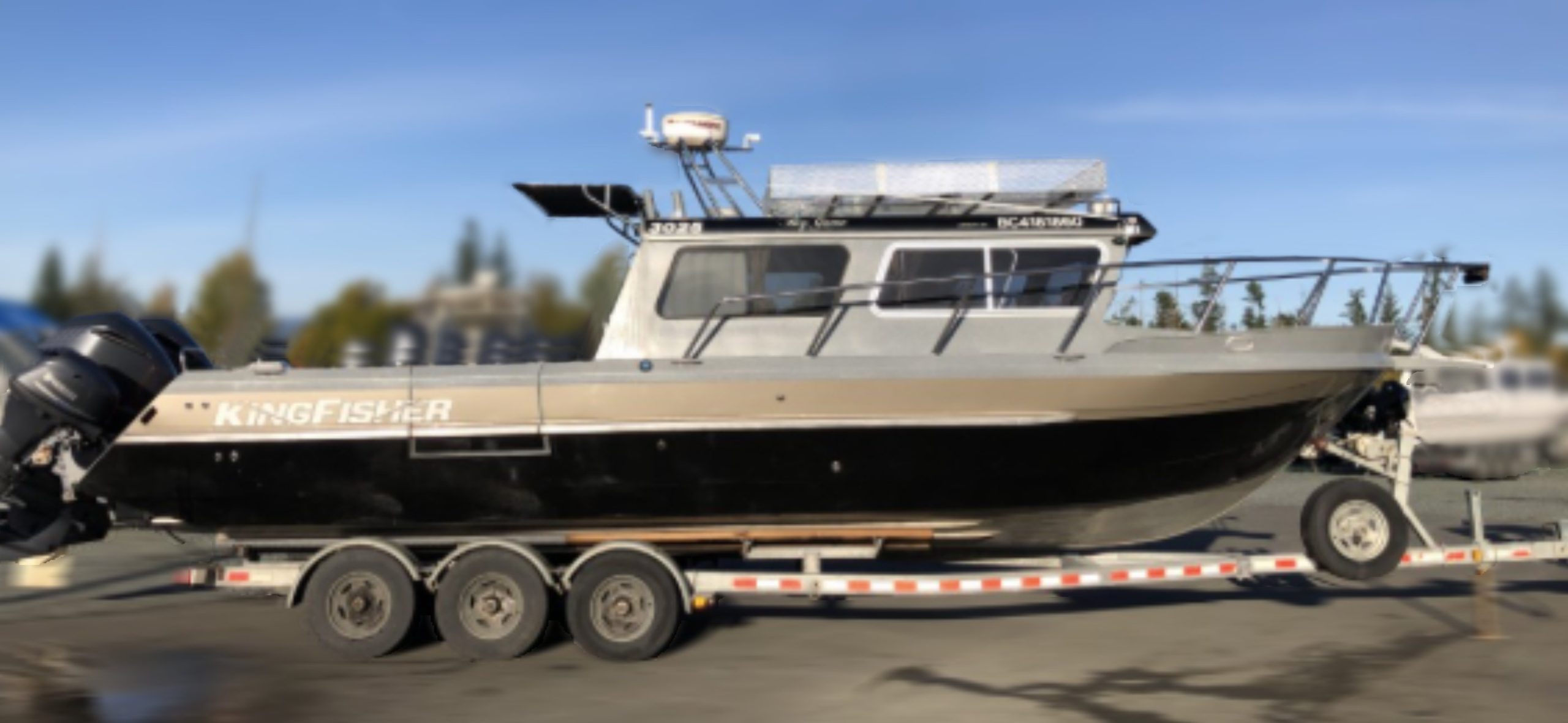 A 2014 / 3025 Kingfisher Destination motorboat in silver and black is secured on a dual-axle trailer in a parking lot, with blue sky and trees visible in the background.