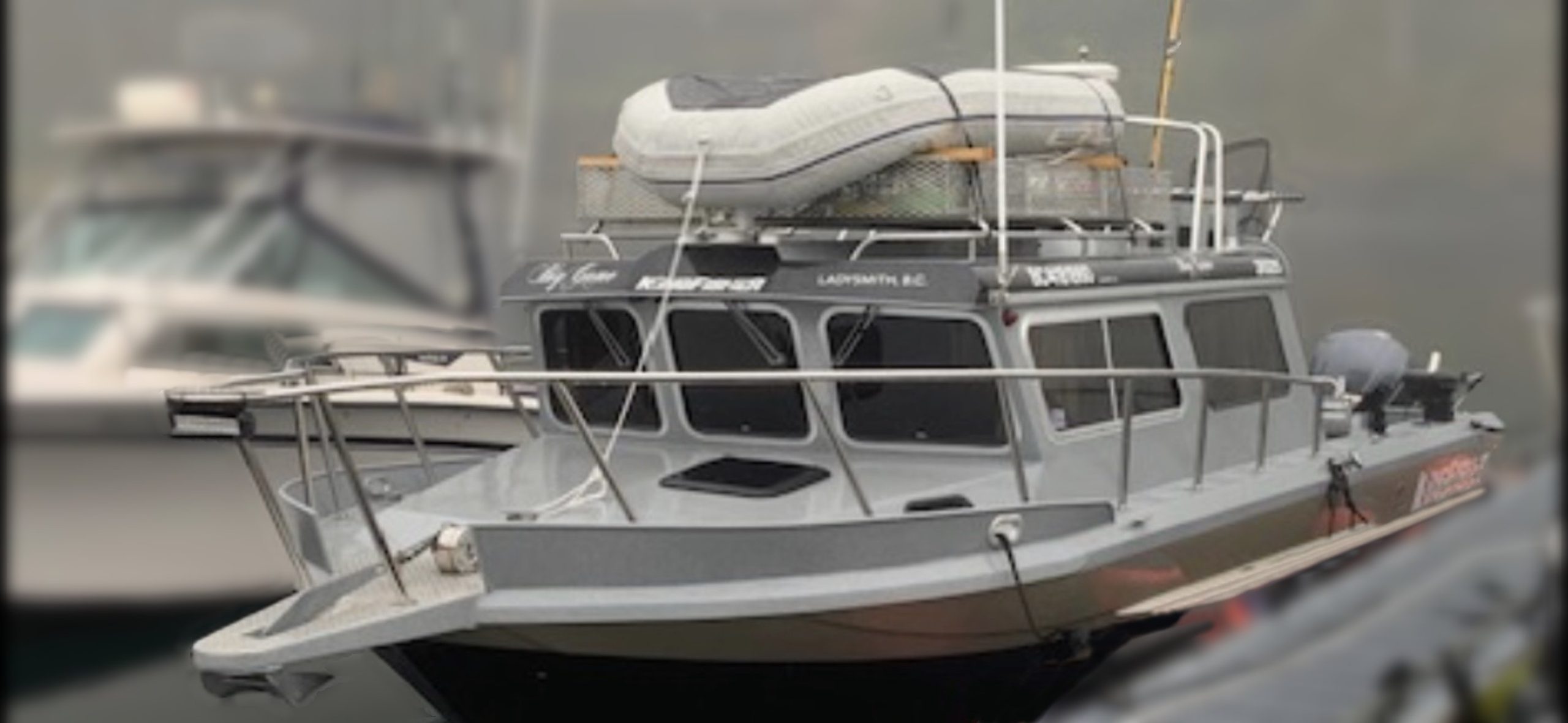 A 2014 / 3025 Kingfisher Destination Pkg motorboat with a silver-gray cabin is docked, featuring a white inflatable dinghy on the roof and front deck safety railings. Another boat can be seen faintly in the background.