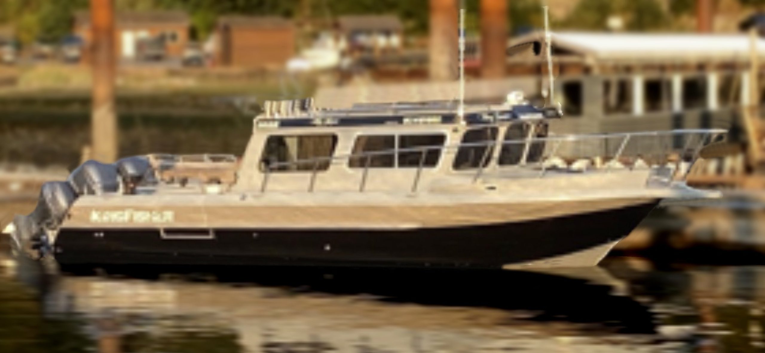 A sleek 2014 / 3025 Kingfisher Destination Pkg motorboat with twin outboards is docked on calm water by a pier, with blurred buildings and trees in the background—ideal for your next adventure.