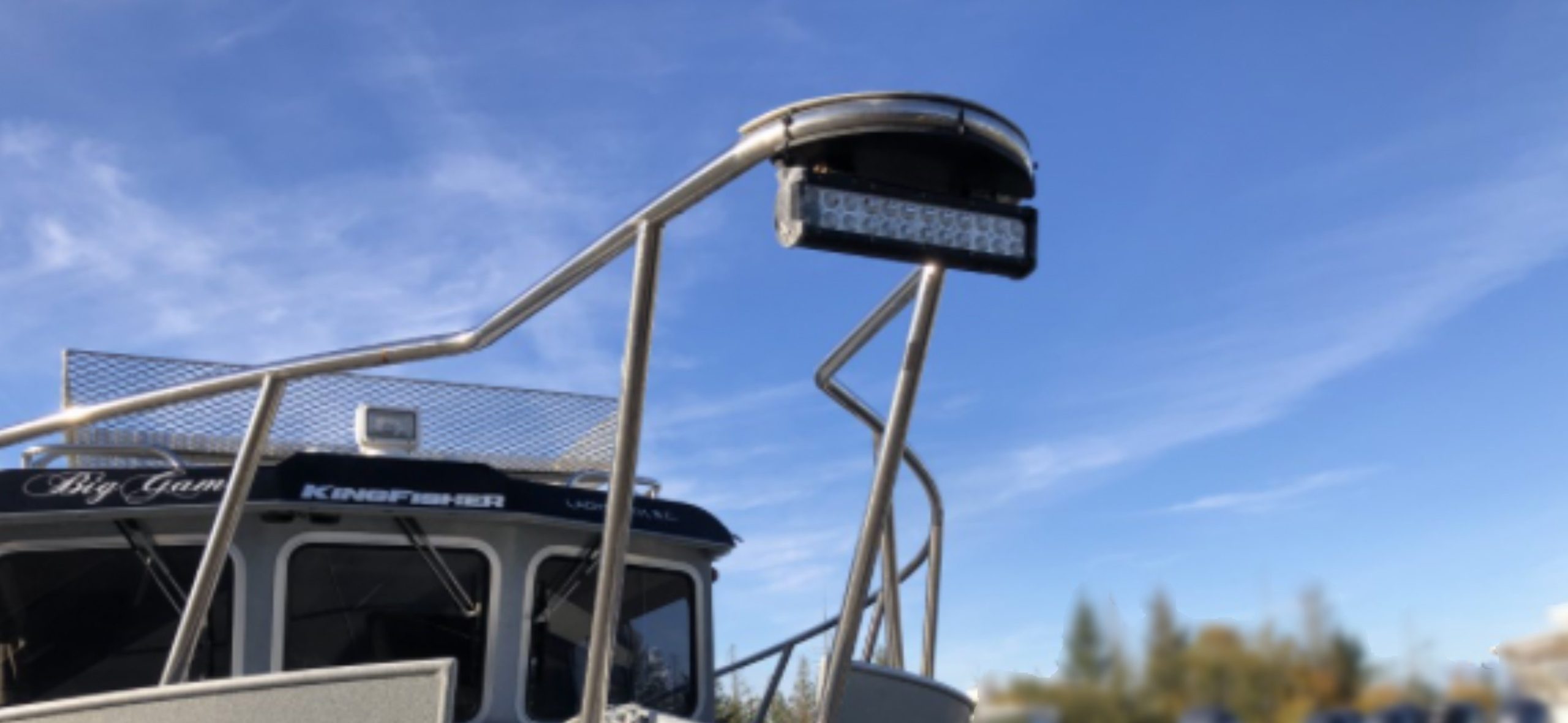 A close-up of a mounted LED floodlight on a metal frame above a boat roof under a clear blue sky. The boat's name "Big Game" and product "2014 / 3025 Kingfisher Destination" are partially visible.