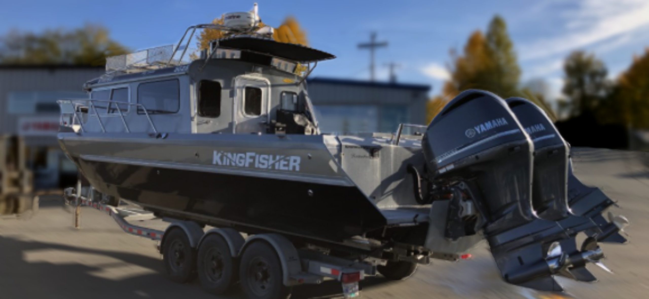 A 2014 / 3025 Kingfisher Destination with twin Yamaha outboards is on a trailer in a parking lot, with a building and trees blurred in the background.