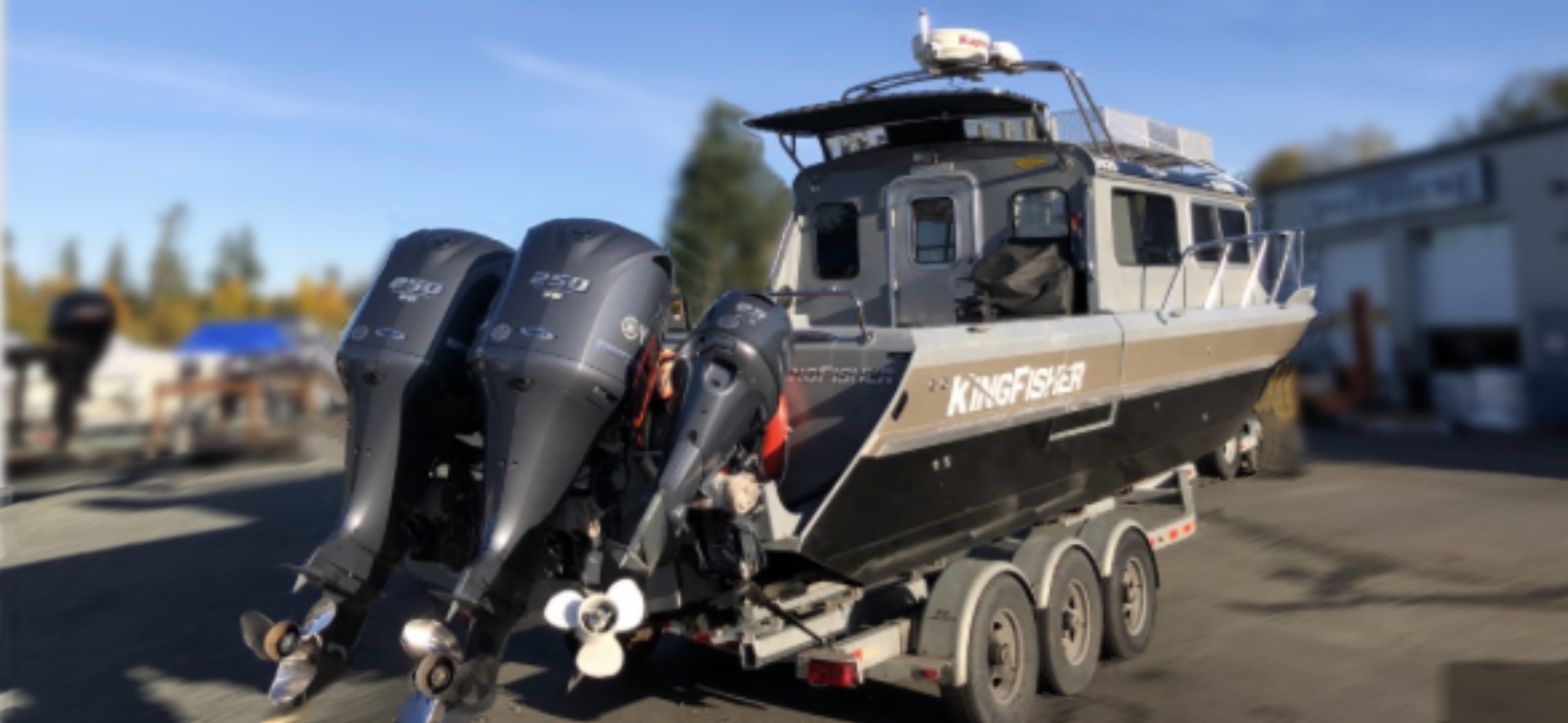 A 2014 / 3025 Kingfisher Destination with twin Yamaha 250 outboards rests on a triple-axle trailer in a parking lot, surrounded by buildings and trees.