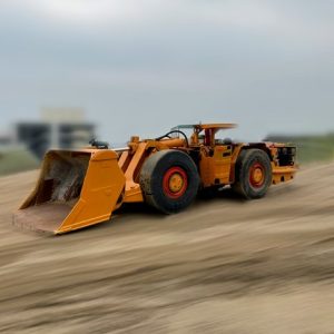 A 1989 Wagner ST-6C Scooptram mining loader with large wheels and a front bucket is parked on dirt, with a blurred sky and buildings in the background.