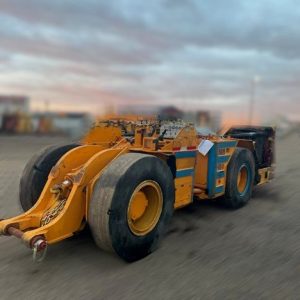 A 2016 RDH 400D Muckmaster Scooptram with oversized rear tires and a low-profile yellow body is parked on gravel under a cloudy sky, with the background softly blurred.