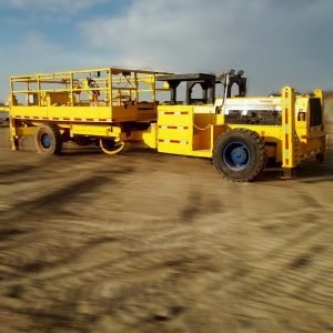 A large yellow mining utility vehicle with a flatbed and safety railings is parked on a dirt surface under a partly cloudy sky. The vehicle has rugged tires and blue wheel rims.