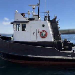 A 1967 / 40FT Mandley Tug in black and white, featuring a red lifebuoy on its side, is docked on calm blue water beneath a clear sky.