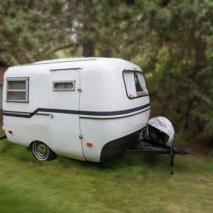 A small, white 1976 Boler 1300 vintage trailer with black trim is parked on grass near trees and a white building with a red roof. It features two small windows and a covered front hitch.