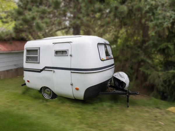 A small, white 1976 Boler 1300 vintage trailer with black trim is parked on grass near trees and a white building with a red roof. It features two small windows and a covered front hitch.