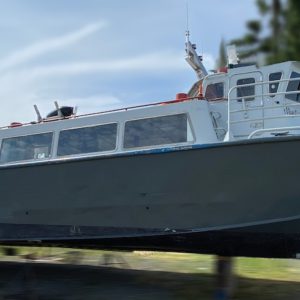 A large, dark-gray 1982 / 50Ft West Coast Passenger Ferry - Crew Vessel is propped up on supports on dry land, with trees and sky in the background. The vessel features side windows and roof equipment.