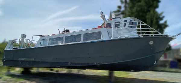 A large, dark-gray 1982 / 50Ft West Coast Passenger Ferry - Crew Vessel is propped up on supports on dry land, with trees and sky in the background. The vessel features side windows and roof equipment.