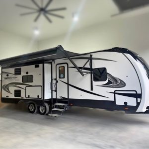 A 2021 Grand Design Reflection 297RSTS travel trailer in white and black, featuring an extended awning, is parked in a spacious, well-lit garage with concrete floors and high ceilings. Steps lead up to the main door.