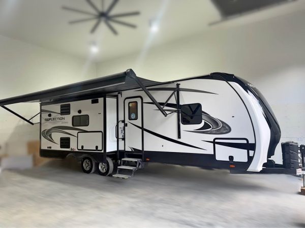 A 2021 Grand Design Reflection 297RSTS travel trailer in white and black, featuring an extended awning, is parked in a spacious, well-lit garage with concrete floors and high ceilings. Steps lead up to the main door.