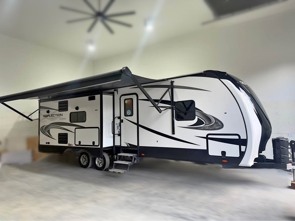 A 2021 Grand Design Reflection 297RSTS travel trailer in white and black, featuring an extended awning, is parked in a spacious, well-lit garage with concrete floors and high ceilings. Steps lead up to the main door.