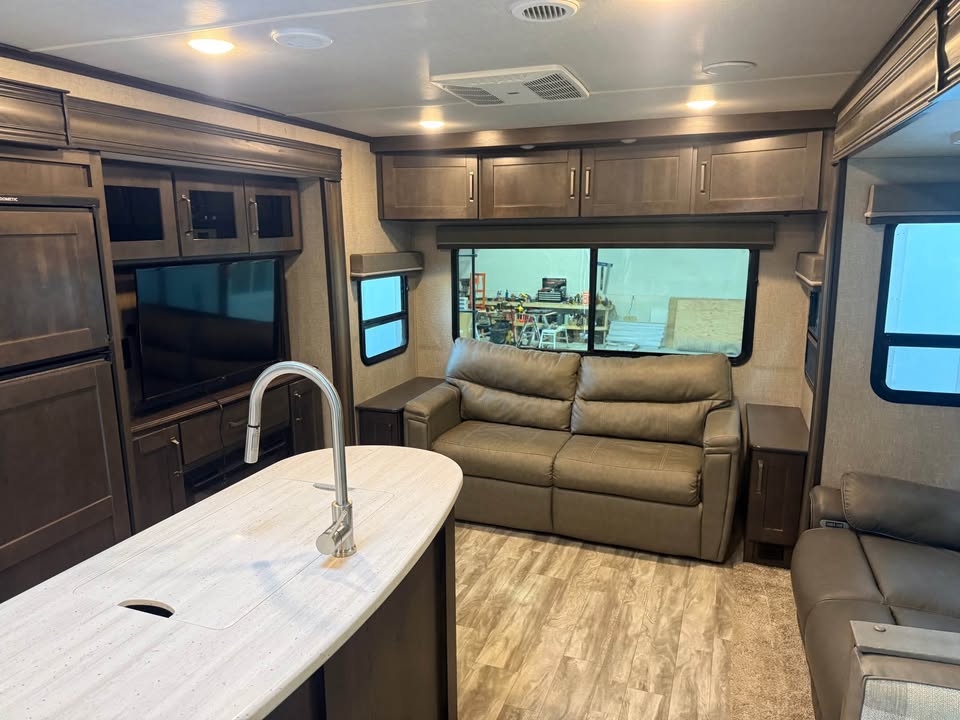 The 2021 Grad Design Reflection 297RSTS features a modern RV interior with dark wood cabinets, beige sofa, large windows, TV, island kitchen with sink, light wood flooring, and neutral decor for a cozy, spacious feel.