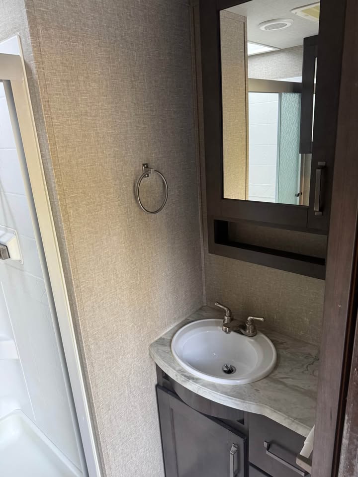The small bathroom in the 2021 Grand Design Reflection 297RSTS includes a marble-look countertop, sink with dual faucets, dark wood cabinet, mirrored medicine cabinet above, wall-mounted towel ring, and a shower stall visible on the left.