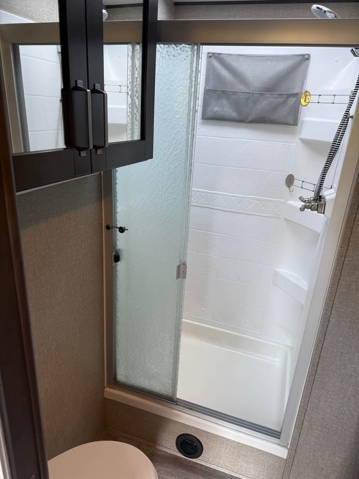 The 2021 Grad Design Reflection 297RSTS features a small bathroom with a frosted glass shower door, white enclosure with shelves, handheld showerhead, wall storage pouch, and mirrored cabinet above the toilet for extra reflection.