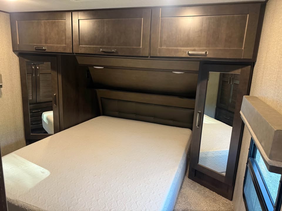 A neatly made bed with a white mattress in the 2021 Grand Design Reflection 297RSTS, featuring dark wood cabinets, mirrored closet doors, a shaded window, and beige carpeted flooring.