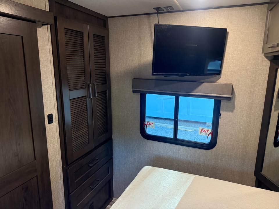 A cozy bedroom with a wooden closet, drawers, wall-mounted TV above a window with exit signs, and a bed with white quilt—perfect for relaxing or working on the 2021 Grad Design Reflection 297RSTS project.