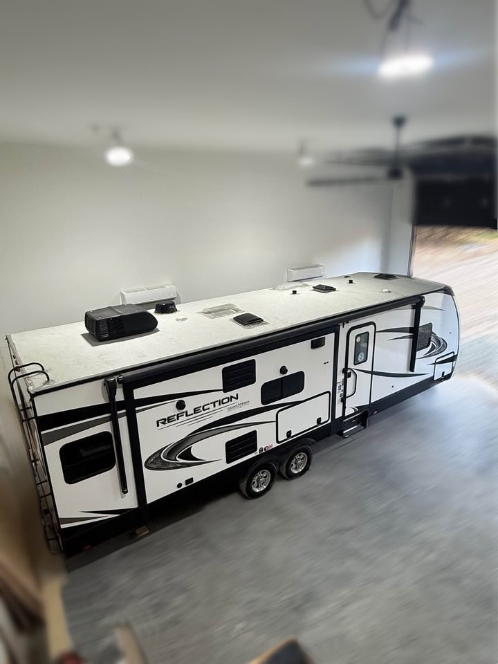 A white 2021 Grad Design Reflection 297RSTS travel trailer with black accents and multiple windows is parked in a spacious, well-lit garage with a concrete floor, as seen from an elevated angle.