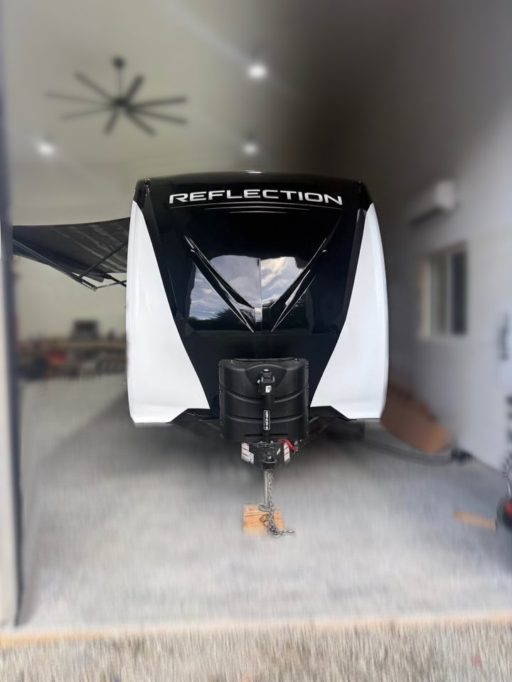 A 2021 Grad Design Reflection 297RSTS in black and white with "REFLECTION" on the front is parked inside a large garage featuring white walls, a ceiling fan, and various items visible in the background.