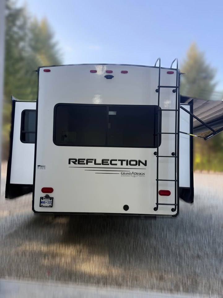 A 2021 Grad Design Reflection 297RSTS RV is shown from the rear with slide-outs extended and a partially open awning, parked on gravel with trees in the background.