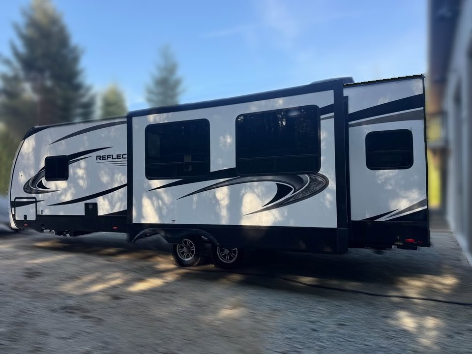A white and black 2021 Grad Design Reflection 297RSTS travel trailer RV with large windows and bold graphics is parked on gravel outdoors, with trees and a building blurred in the background.