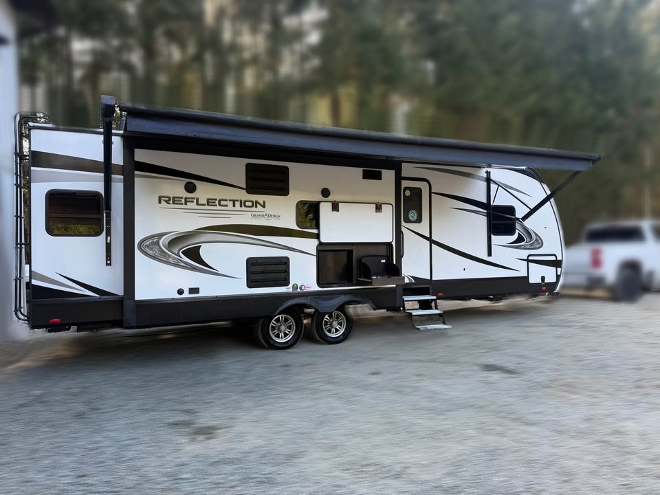 A 2021 Grad Design Reflection 297RSTS, a large white and black travel trailer, is parked outdoors with its awning extended. Steps lead to the door, with trees and a vehicle visible in the background.